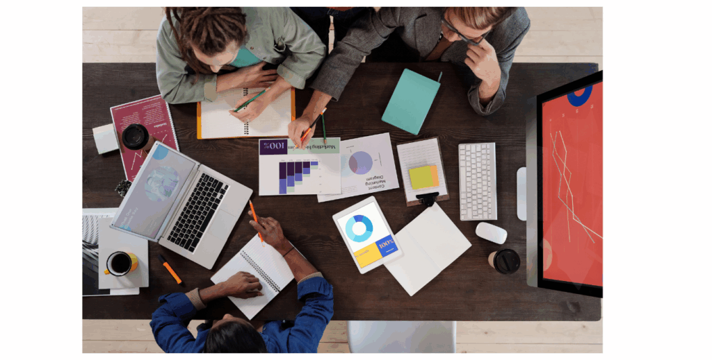3 people at a desk with a computer, laptop and papers
